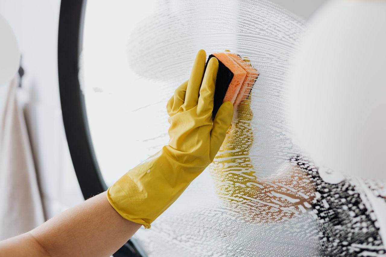 Personne anonyme portant des gants en caoutchouc jaunes utilisant une simple éponge pour nettoyer un miroir dans une salle de bain